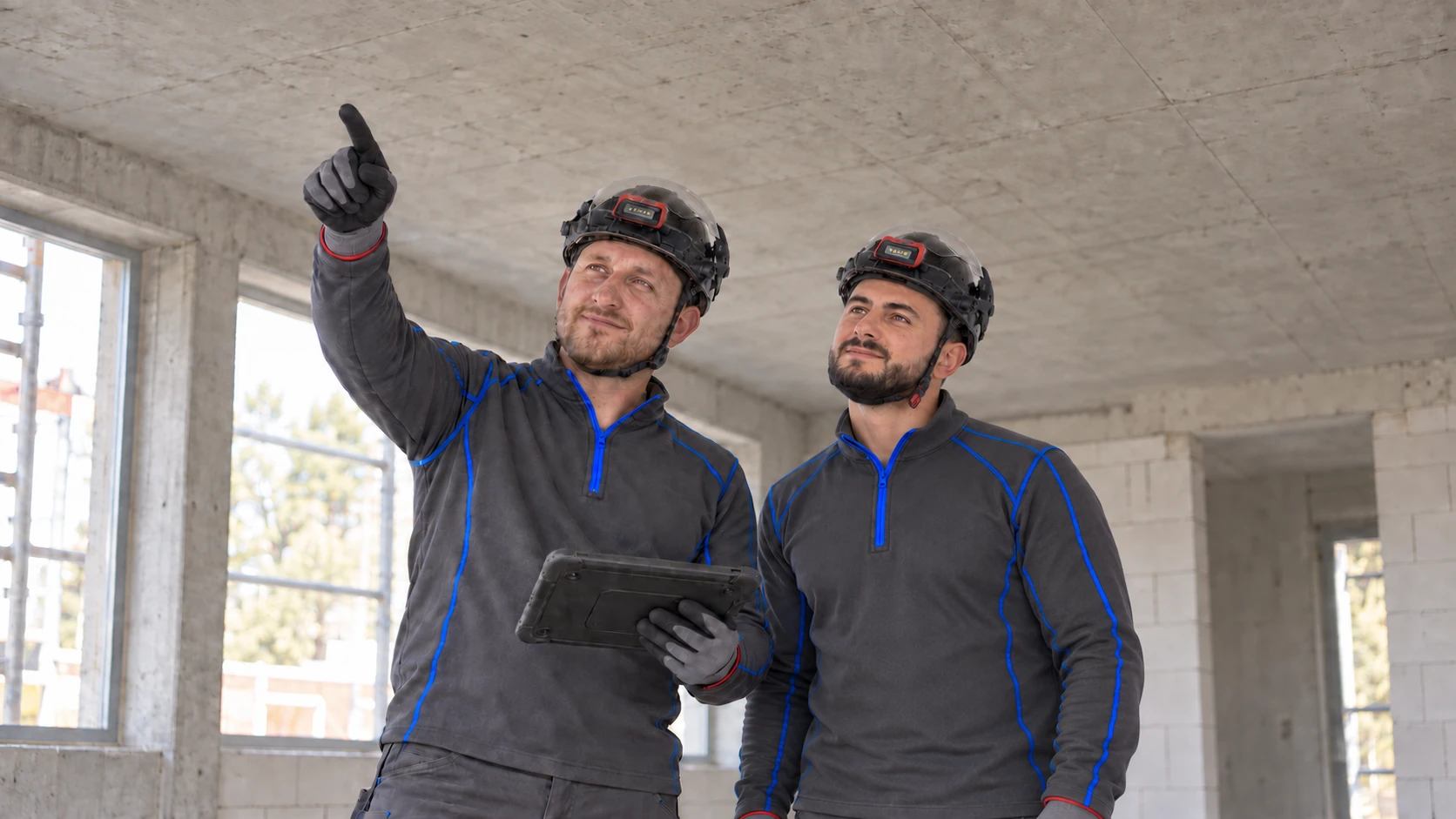 KI-generierte Rohbaubesprechung: Bujar Osmanaj hält ein Tablet und zeigt zur freien Betondecke. Bujar Avdijaj folgt mit Helm seinem Fingerzeig.